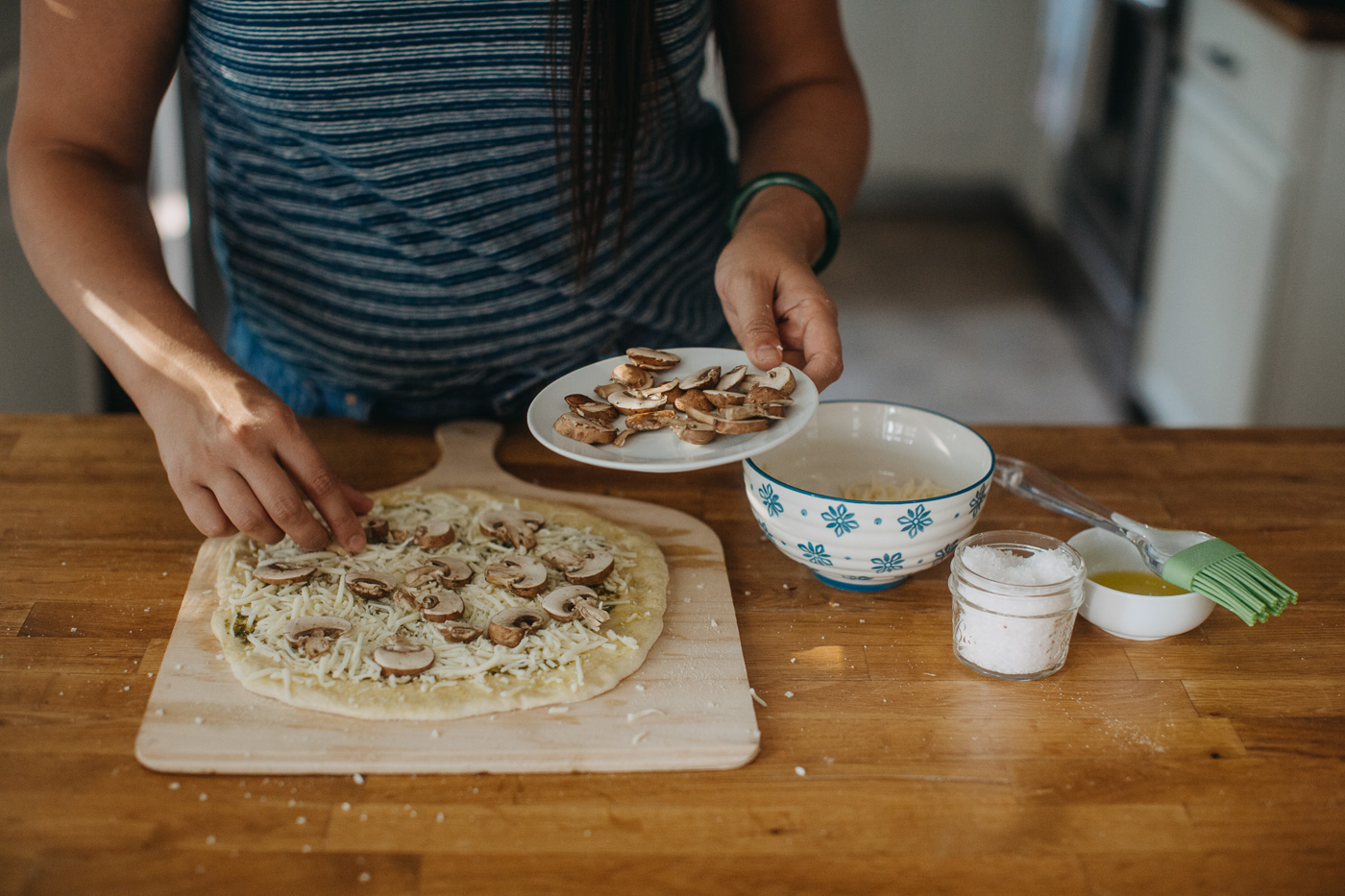 woman in blue shirt showing how to make homemade pizza for pizza night by adding thinly sliced mushrooms to homemade pizza.