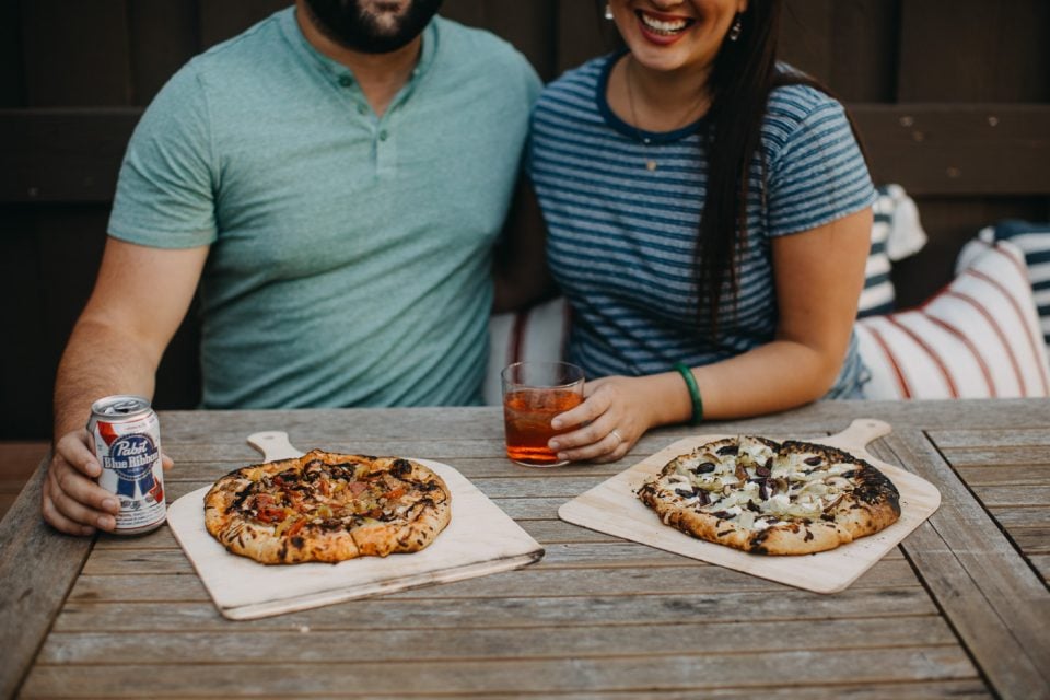 man & woman sitting at patio table enjoying homemade pizzas & drinks for pizza night.