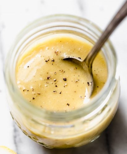All purpose vinaigrette in jar set on white marble backdrop. Spoon coming out of jar. Cracked black pepper on surface of salad dressing. Lemons in the background.