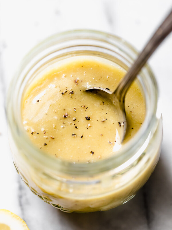 All purpose vinaigrette in jar set on white marble backdrop. Spoon coming out of jar. Cracked black pepper on surface of salad dressing. Lemons in the background.