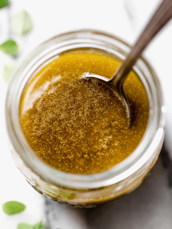 Easy Greek Dressing in small jar on white marble surface. Patina spoon in jar, small green fresh oregano leaves scattered around.