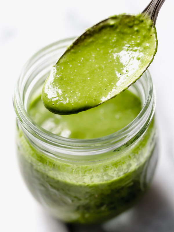 Green Goddess dressing in small jar with spoon drizzling dressing into jar. Jar placed on white marble surface with fresh chives & basil leaves around it.