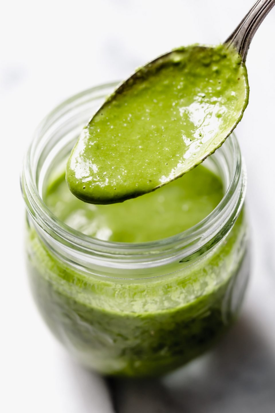 Green Goddess dressing in small jar with spoon drizzling dressing into jar. Jar placed on white marble surface with fresh chives & basil leaves around it.