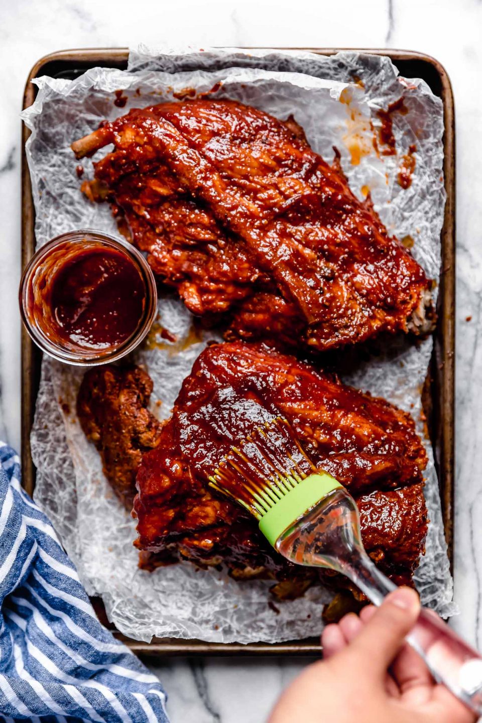 Baby back ribs sitting on a parchment-paper lined baking sheet. Woman's hand is brushing the ribs with BBQ sauce. The baking sheet is on a white marble surface with a striped blue linen nearby.