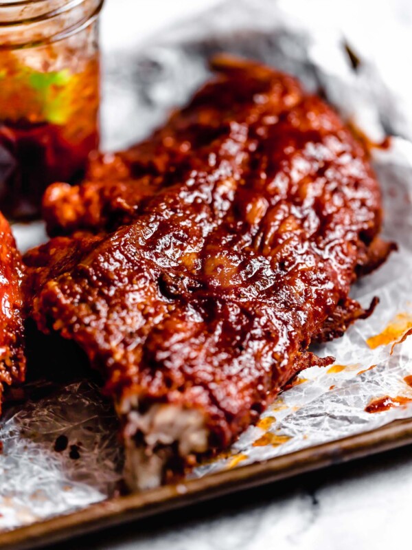 Close up of baby back ribs sitting on a parchment-lined baking sheet with a jar of BBQ sauce in the background.