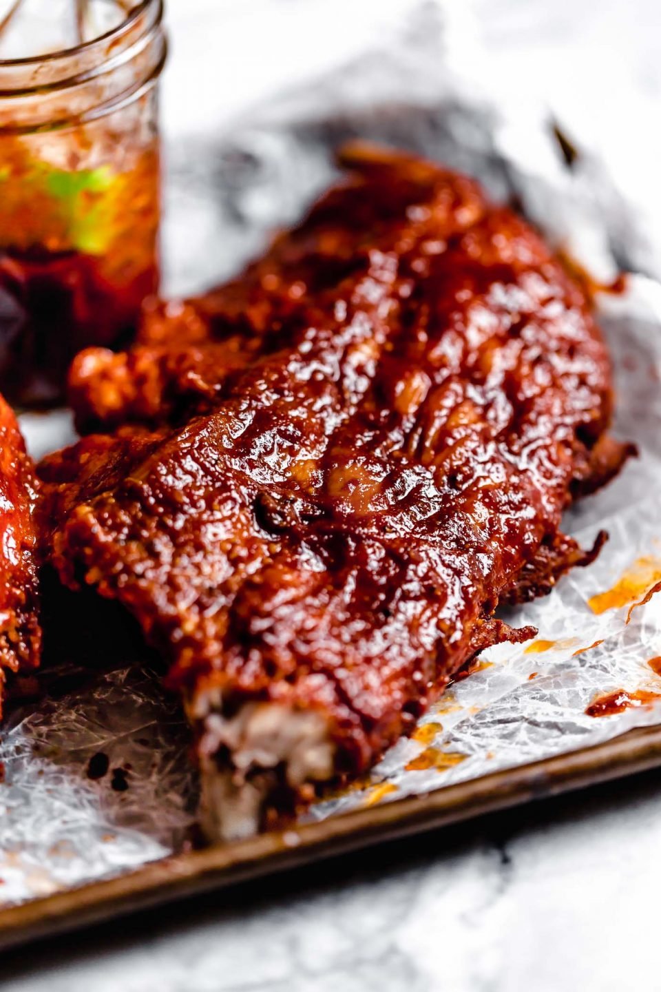 Close up of baby back ribs sitting on a parchment-lined baking sheet with a jar of BBQ sauce in the background.