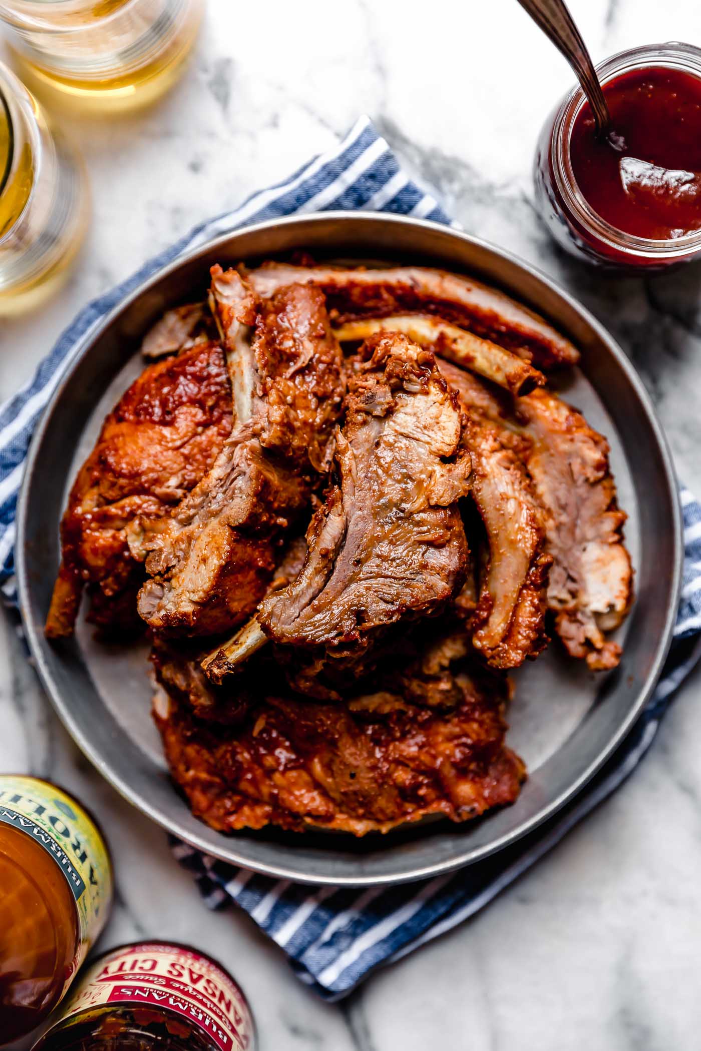 Baby back ribs served in a round metal dish, with BBQ sauce & beer. The dish is sitting on a blue & white striped linen atop a white marble surface. 