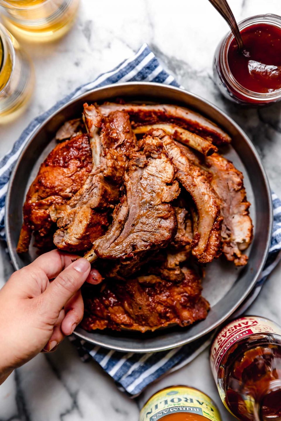 A woman's hand is picking up a single baby back ribs served with other baby back ribs in a round metal dish, with BBQ sauce & beer. The dish is sitting on a blue & white striped linen atop a white marble surface. 