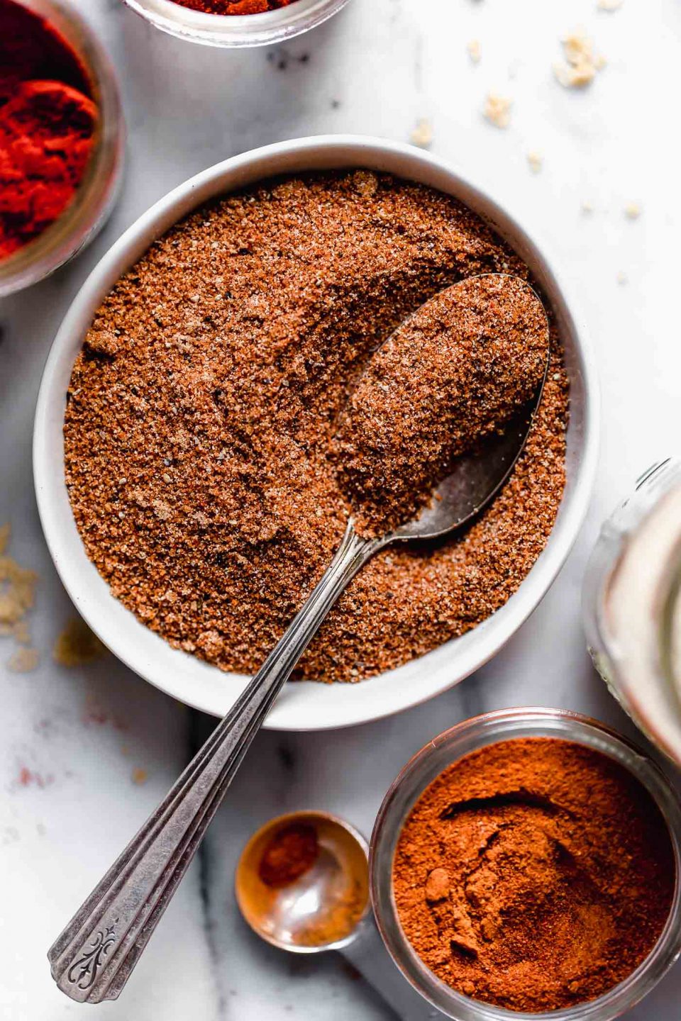 Mixed dry rub for baby back ribs shown in a small white bowl with a patina spoon. The dry rub bowl is shown on a marble surface, next to a small measuring spoon & some larger jars of spices.