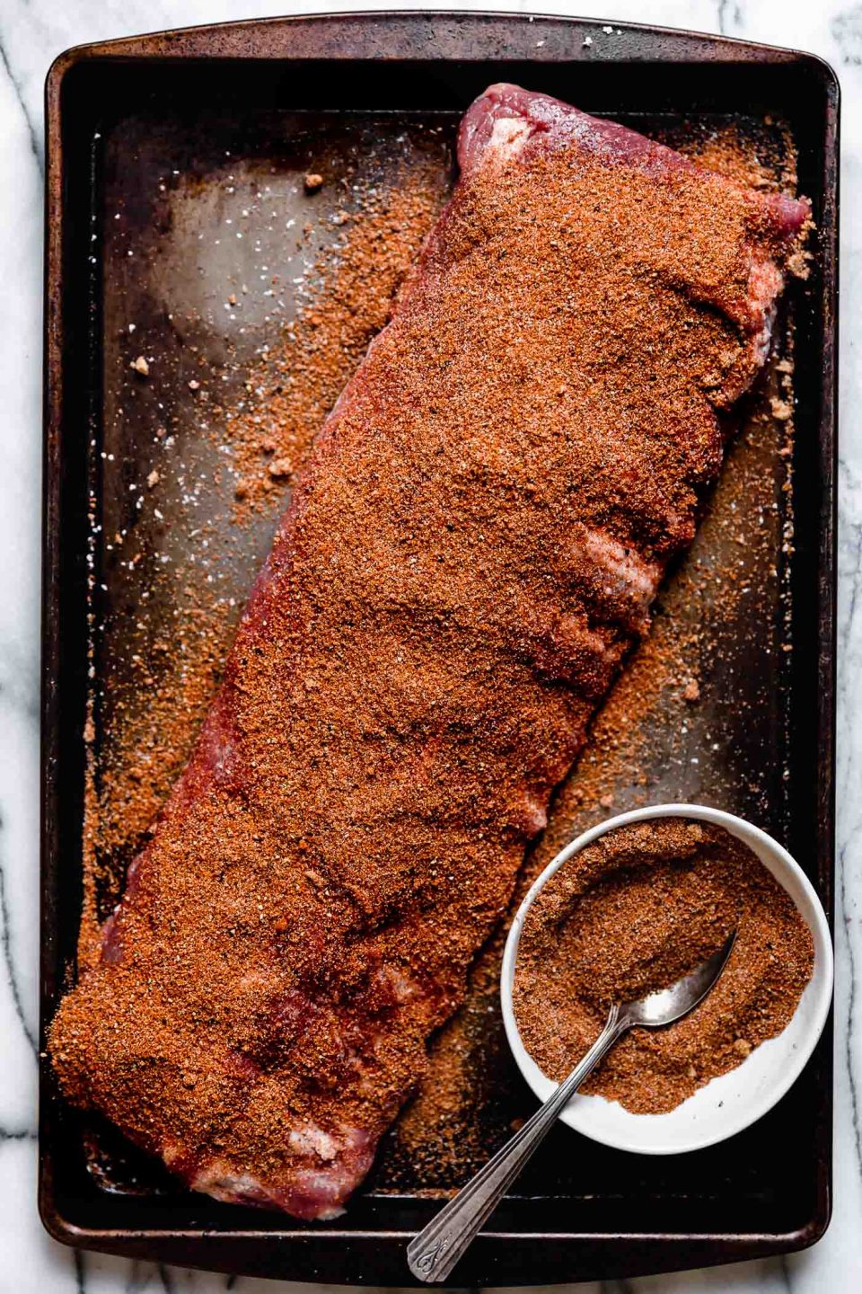A rack of ribs, seasoned generously with dry rub for baby back ribs, on a patina baking sheet on a white marble surface.