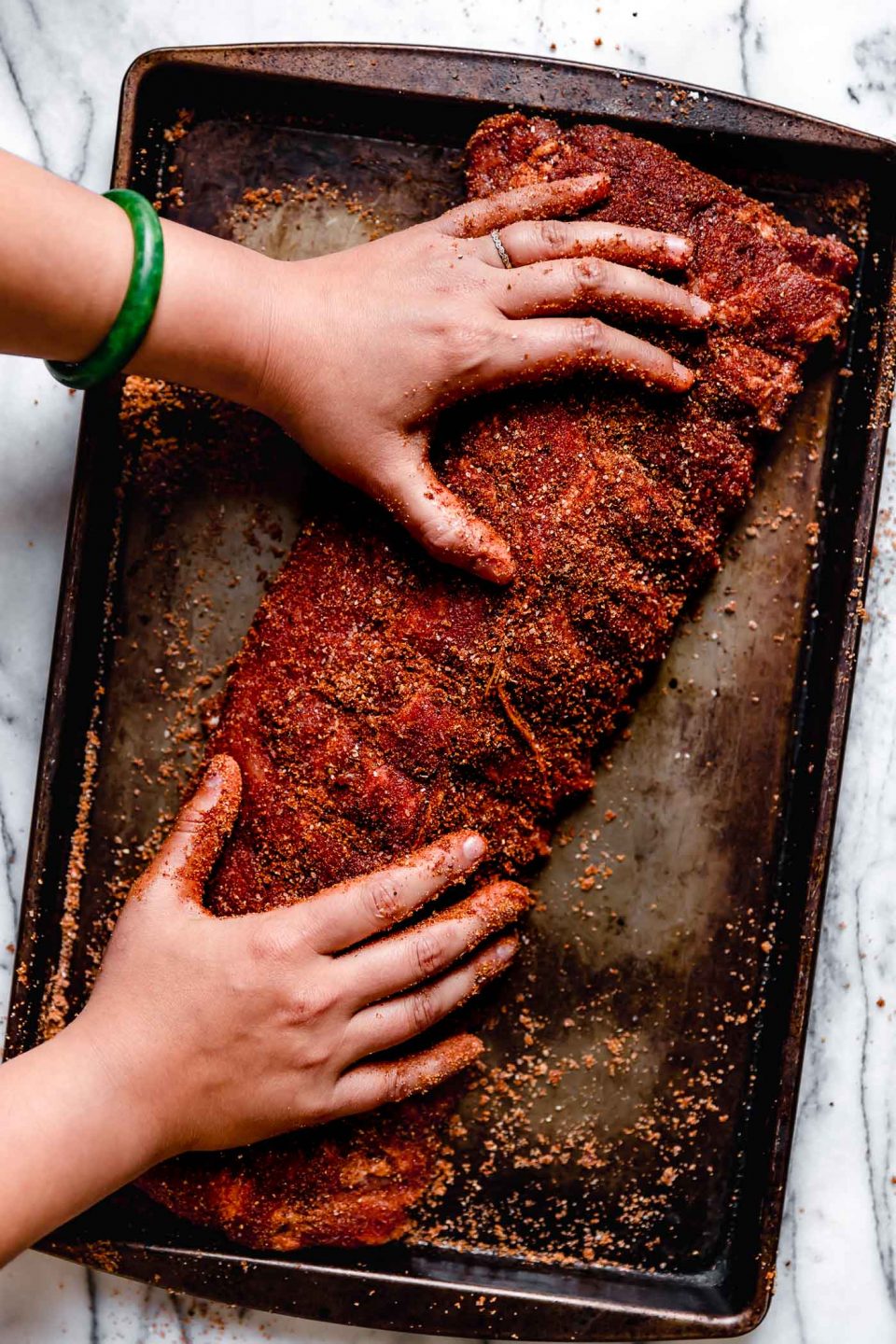 A rack of ribs, seasoned generously with dry rub for baby back ribs, on a patina baking sheet on a white marble surface. Woman's hands reaching into the frame to rub the dry rub into the ribs.