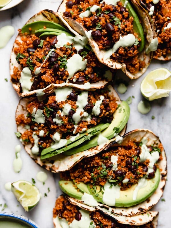 Quinoa black bean tacos arranged on a marble board, topped with cilantro lime crema sauce and chopped cilantro. There are a few lime wedges surrounding the tacos, along with a small blue bowl with the cilantro lime crema.