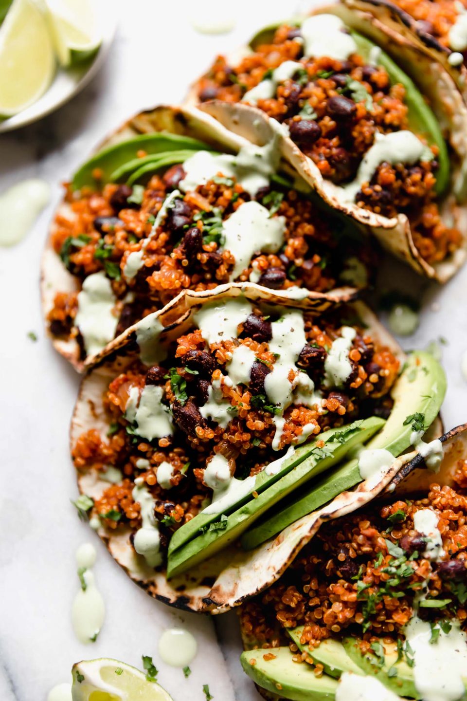 Quinoa Black Bean Tacos arranged on a marble board, topped with cilantro lime crema sauce & chopped cilantro. There are a few lime wedges surrounding the tacos, along with a small blue bowl with the cilantro lime crema.