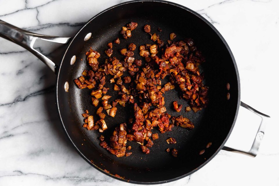 How to make Quinoa Taco Meat - Step 2: Aromatics coating softened onion in black skillet atop a white marble surface.