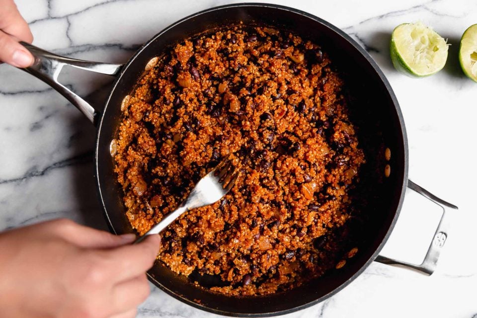 Hand reaching into the photo fluffing cooked quinoa & black bean taco filling (vegetarian taco meat) with a fork. The skillet is placed on a marble surface, with a few spent lime halves surrounding it.