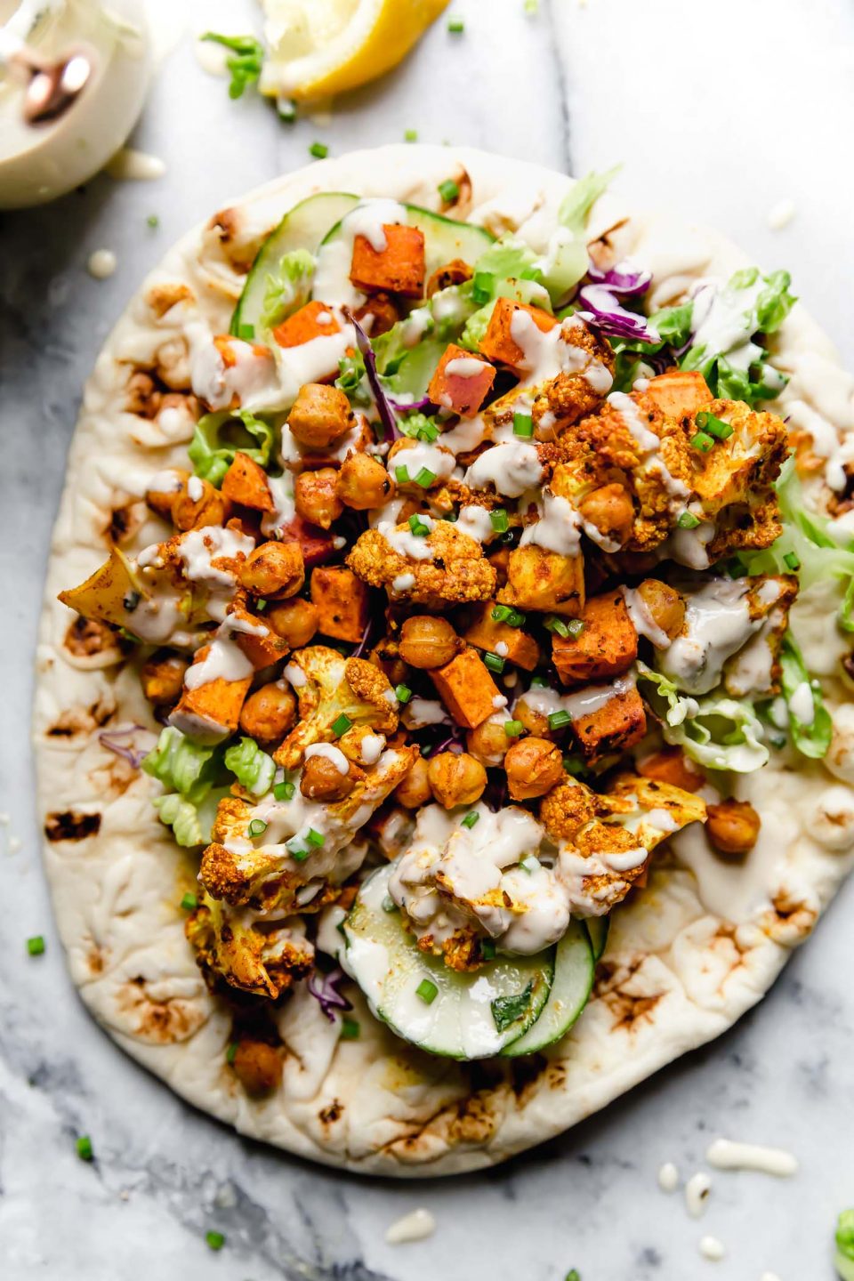 Chickpea & veggie shawarma served on a piece of naan bread. The bread is placed on a white marble surface, alongside lemon wedges & a jar of tahini sauce.