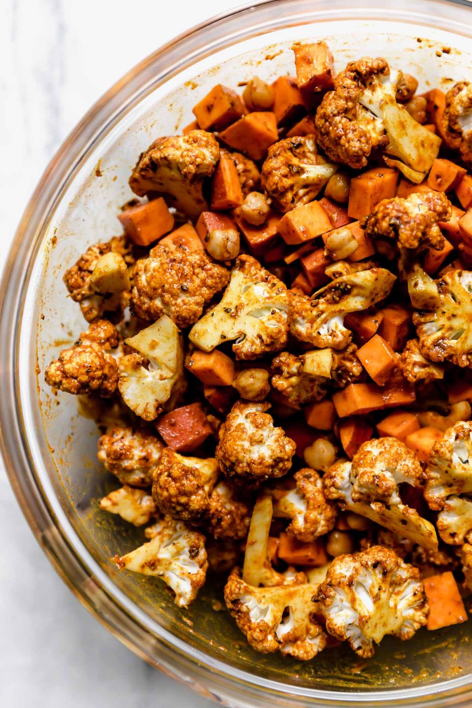 Sweet potato, cauliflower and chickpeas are tossed in shawarma marinade inside of a large glass bowl.