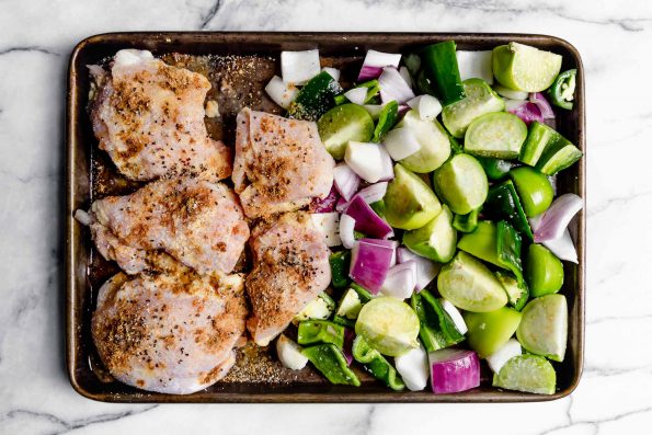 Salsa verde chicken ingredients (chicken thighs, tomatillos, red onion, jalapeno & poblano) on a sheet pan. Veggies are one one side of the pan and the chicken is opposite. Sheet pan is placed on a white marble surface.