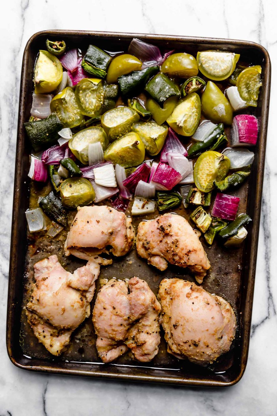 Salsa verde chicken ingredients (chicken thighs, tomatillos, red onion, jalapeno & poblano) on a sheet pan after having been roasted. Roasted veggies are one one side of the pan and the chicken is opposite. Sheet pan is placed on a white marble surface.