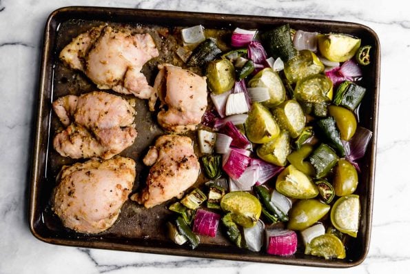Salsa verde chicken ingredients (chicken thighs, tomatillos, red onion, jalapeno & poblano) on a sheet pan after having been roasted. Roasted veggies are one one side of the pan and the chicken is opposite. Sheet pan is placed on a white marble surface.