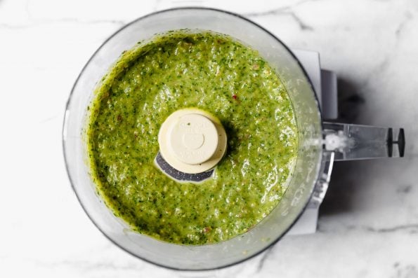 Blended salsa verde in food processor on a white marble surface.