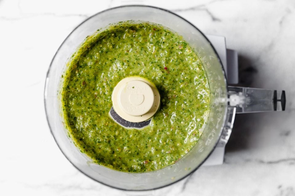 Blended salsa verde in food processor on a white marble surface.