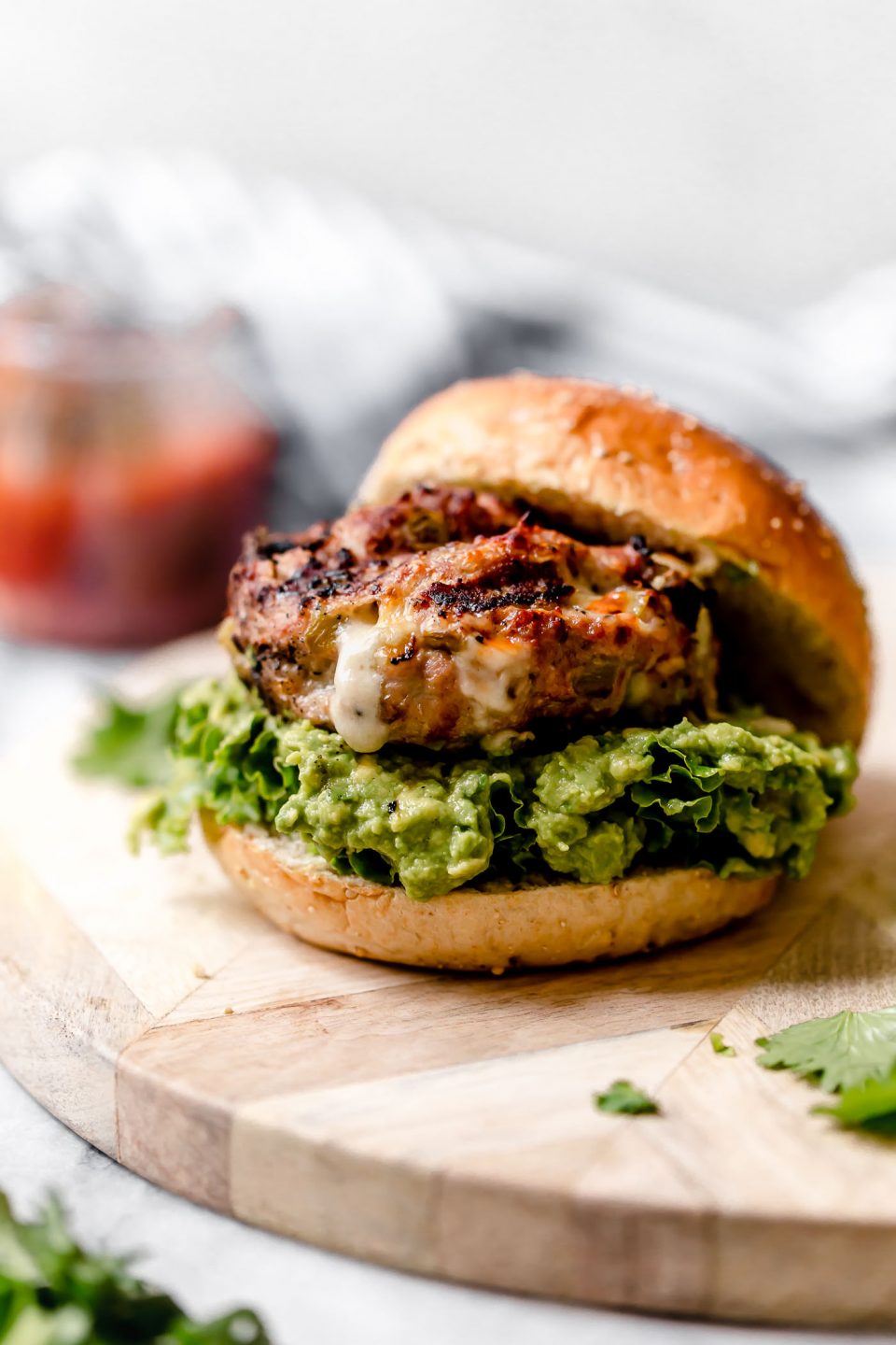 Stuffed Southwest Turkey Burger place on a burger bun with lettuce & guacamole. Burger is sitting on a light wood board, with a jar of salsa behind it.