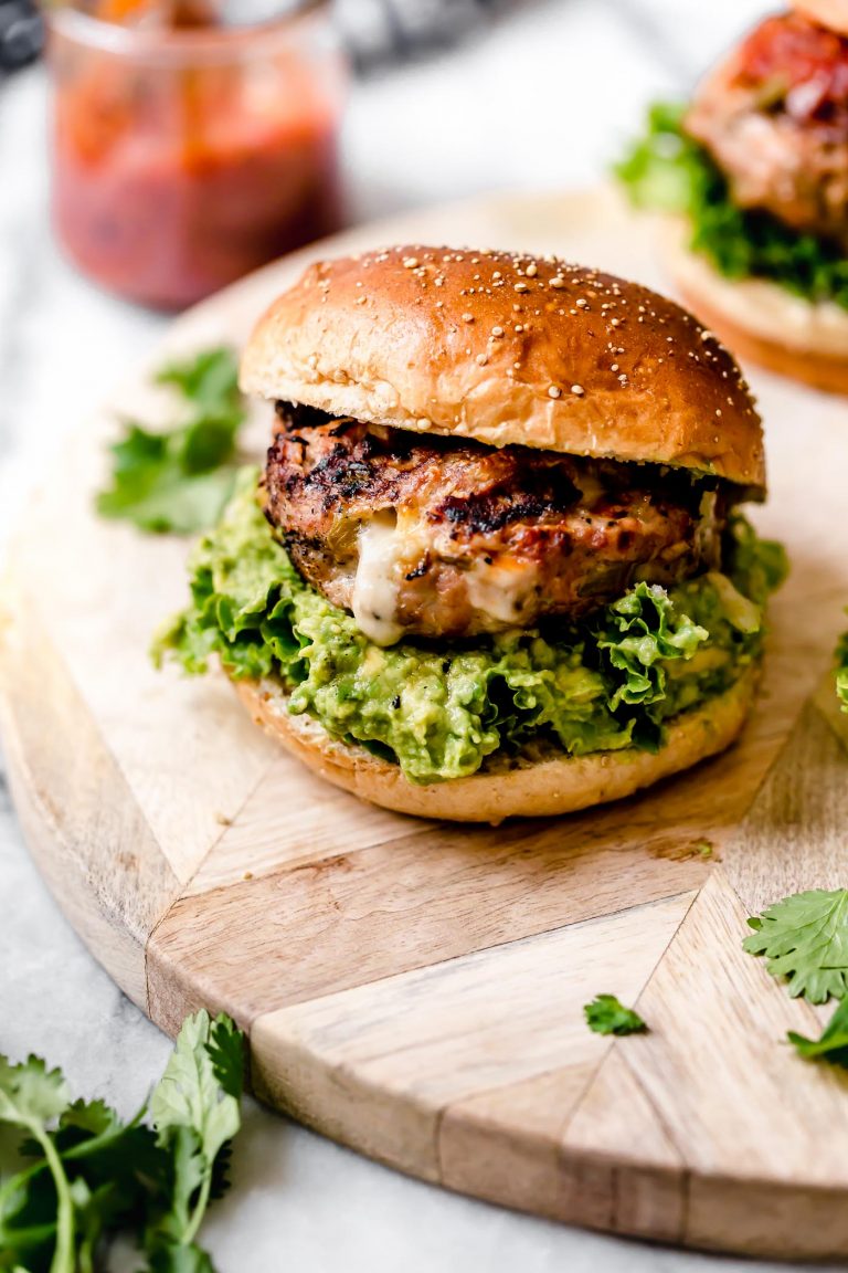 Stuffed Southwest Turkey Burger place on a burger bun with lettuce & guacamole. Burger is sitting on a light wood board, with a jar of salsa behind it.