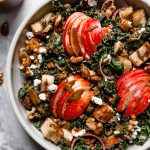 Tossed Autumn Kale salad in a cream ceramic bowl on a white marble surface. The salad is topped with clusters of sliced honeycrisp apples & croutons, & an extra drizzle of maple balsamic vinaigrette.
