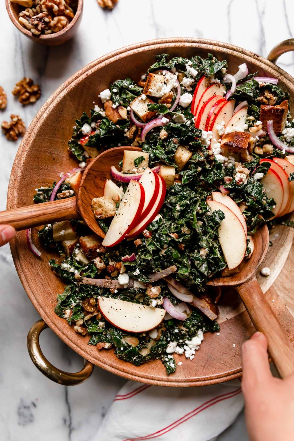 Autumn Kale salad in a large wooden serving bowl. A woman's hands are reaching into the frame with wooden spoons, tossing the salad. The salad is topped with clusters of sliced honeycrisp apples & croutons. The serving bowl is atop a white marble surface, next to honeycrisp apples & whole walnuts.