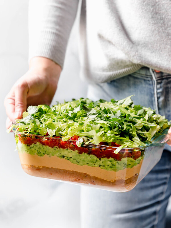 Woman in blue jeans & a gray sweater holding Vegan 7-Layer Dip in front of her.
