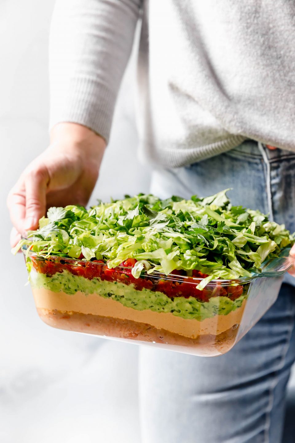 Woman in blue jeans & a gray sweater holding Vegan 7-Layer Dip in front of her.