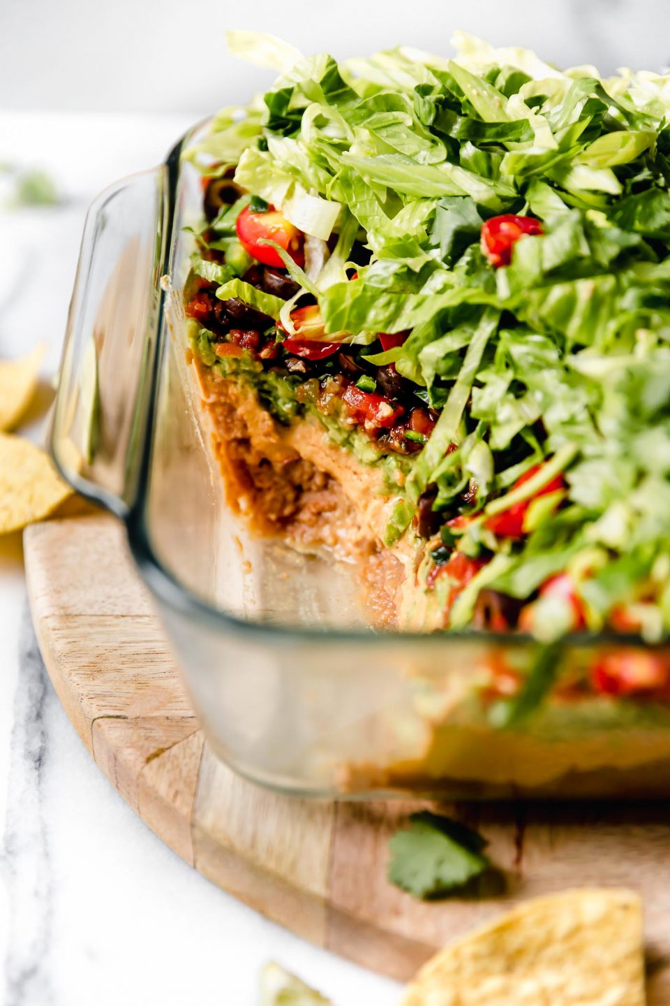 Straight-on photo of this vegan 7-Layer Dip recipe shown in clear Pyrex DEEP baking dish. The dish is sitting on a light wood board, with some of the dip removed from the container.