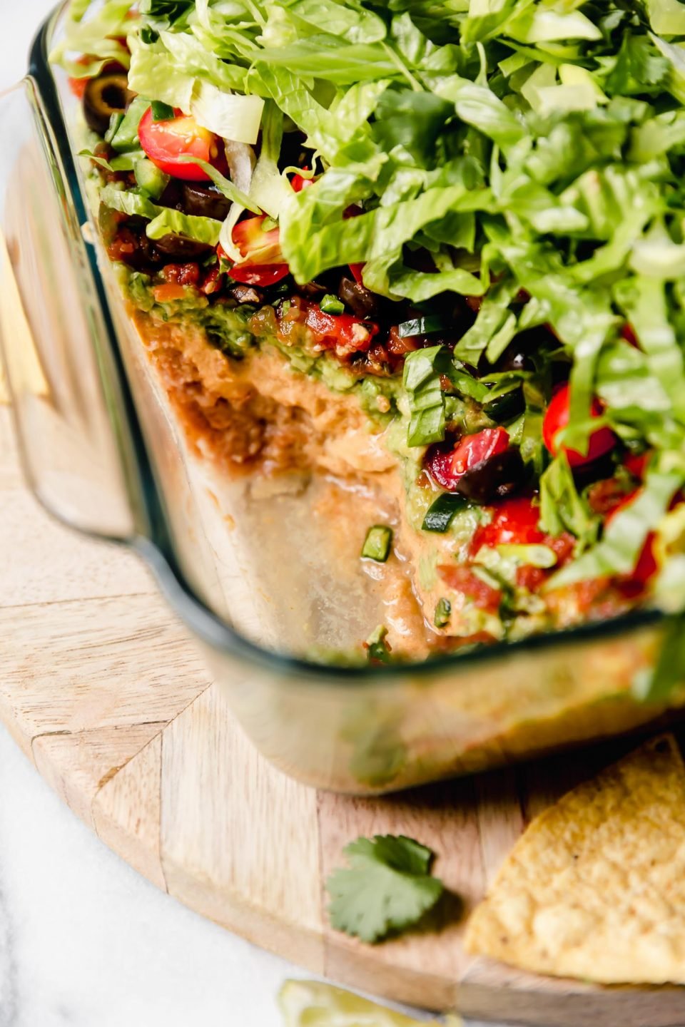 Straight-on photo of this vegan 7-Layer Dip recipe shown in clear Pyrex DEEP baking dish. The dish is sitting on a light wood board, with some of the dip removed from the container.