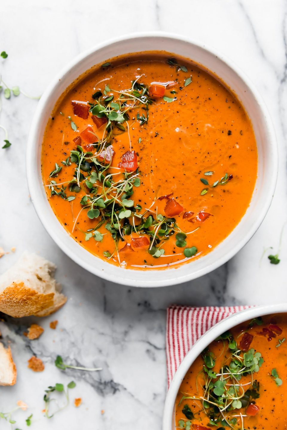 Creamy Roasted Red Pepper soup in a white ceramic bowl, topped with microgreens and cracked black pepper. The bowl is surrounded by some pieces of crusty bread, and a second bowl of soup sitting on a striped red linen napkin. Everything is placed on a white marble surface.
