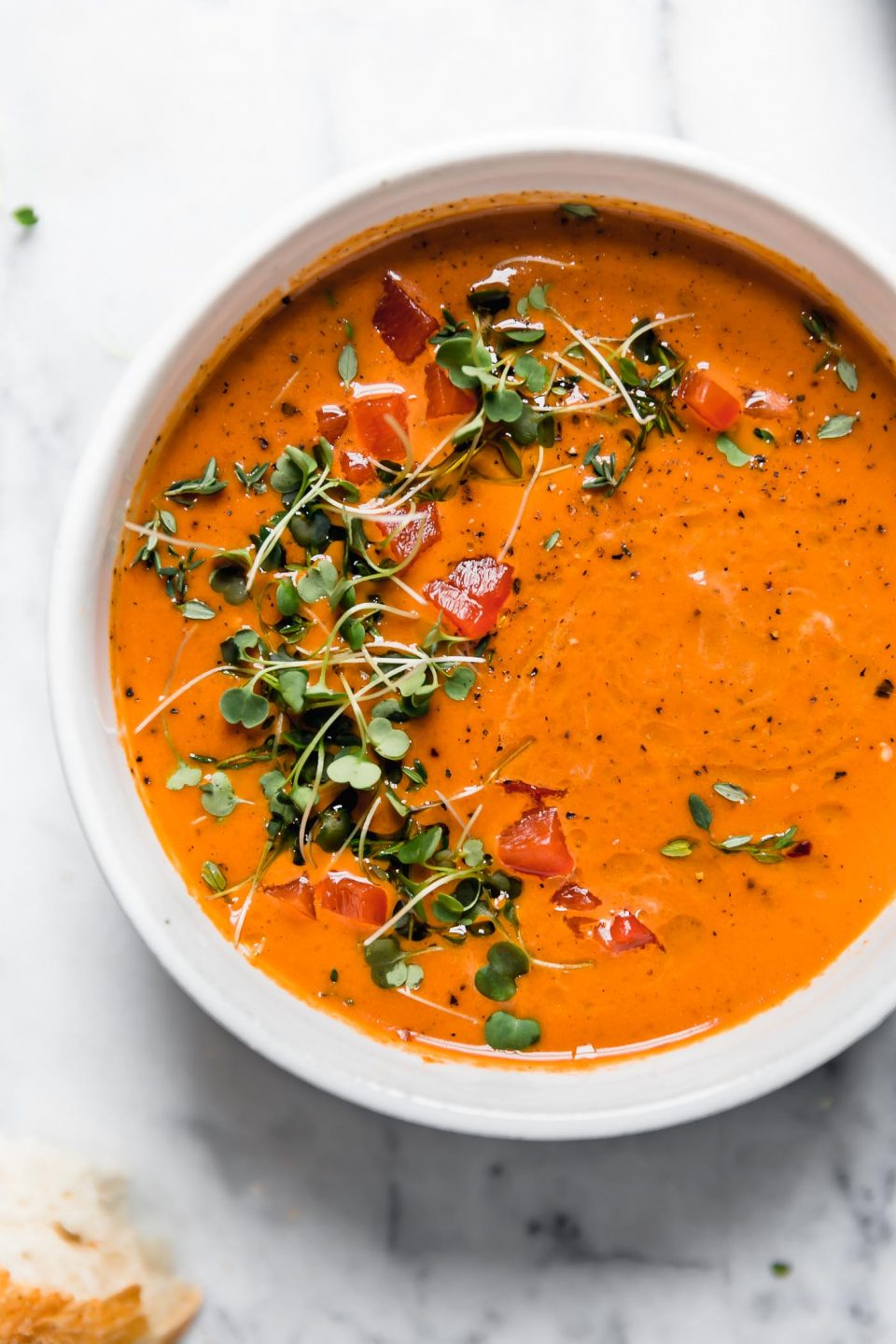 Creamy Roasted Red Pepper soup in a white ceramic bowl, topped with microgreens and cracked black pepper. The bowl is surrounded by some pieces of crusty bread, and a second bowl of soup. They are sitting atop a white marble surface.