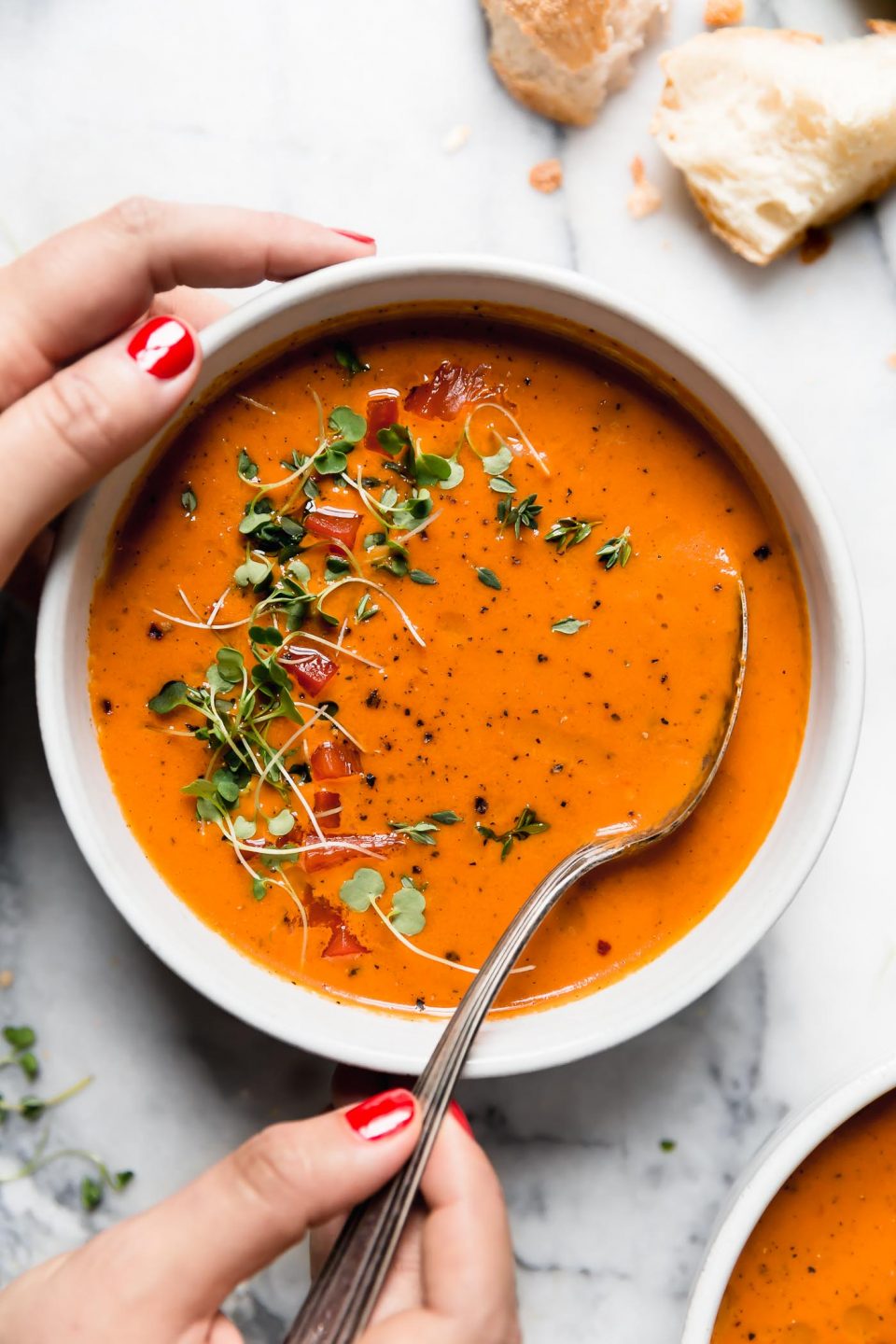 Creamy Roasted Red Pepper soup in a white ceramic bowl, topped with microgreens and cracked black pepper. The bowl is surrounded by some pieces of crusty bread, and a second bowl of soup. They are sitting atop a white marble surface. A woman's manicured hands are reaching in to take a spoonful of soup out of the bowl. 