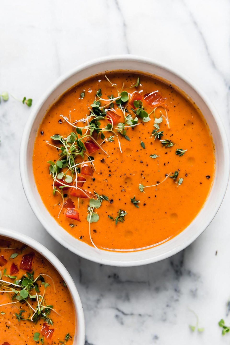 Creamy Roasted Red Pepper soup in a white ceramic bowl, topped with microgreens and cracked black pepper. The bowl is next to a second bowl of soup. They are sitting atop a white marble surface.
