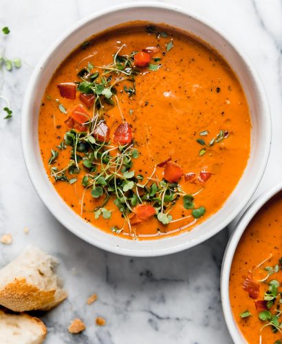 Creamy Roasted Red Pepper soup in a white ceramic bowl, topped with microgreens and cracked black pepper. The bowl is surrounded by some pieces of crusty bread, and a second bowl of soup. They are sitting atop a white marble surface.