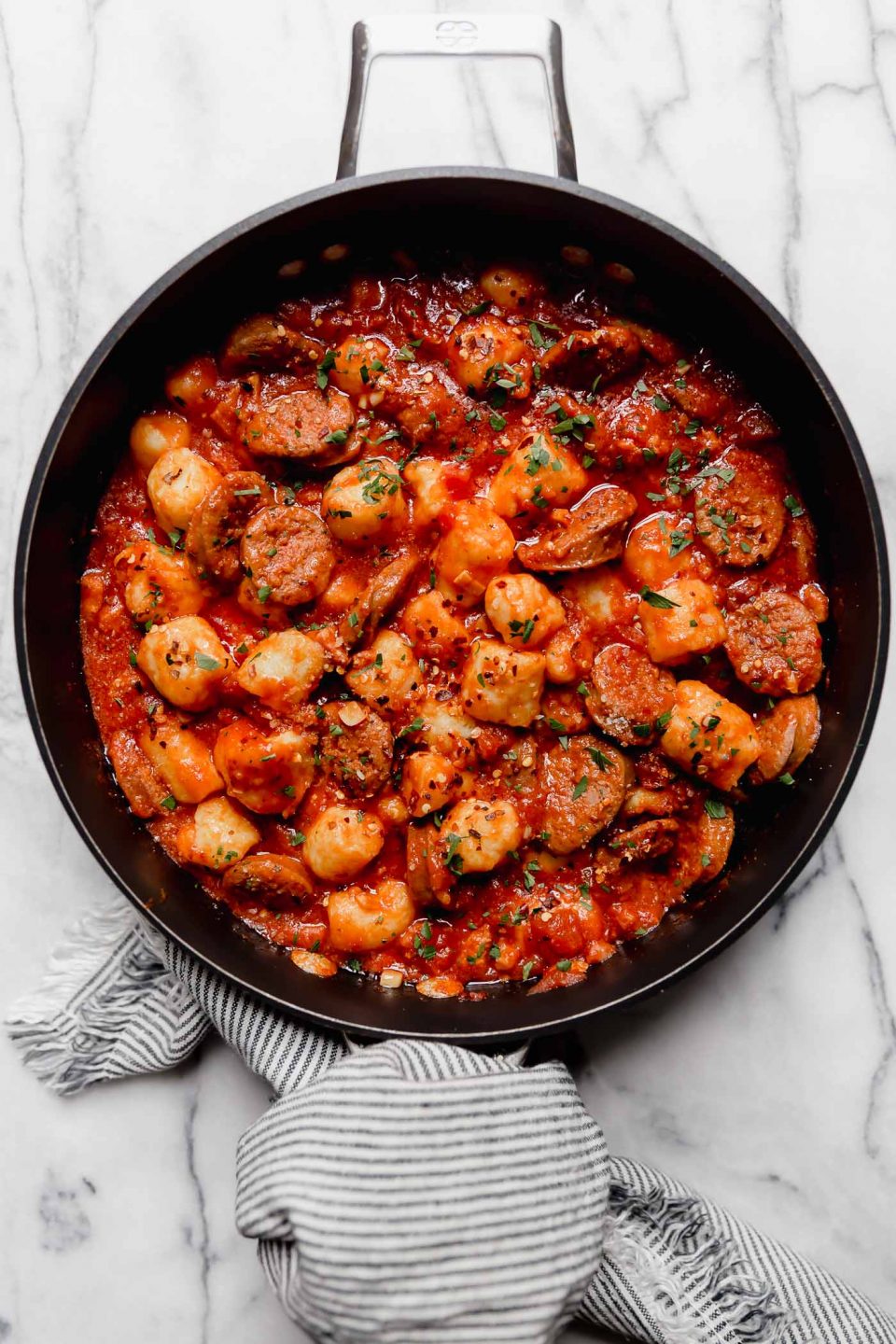 Gnocchi all'Amatriciana, with sliced Italian sausage in a black skillet. A striped linen is wrapped around the skillet handle. The skillet is on a white marble surface.