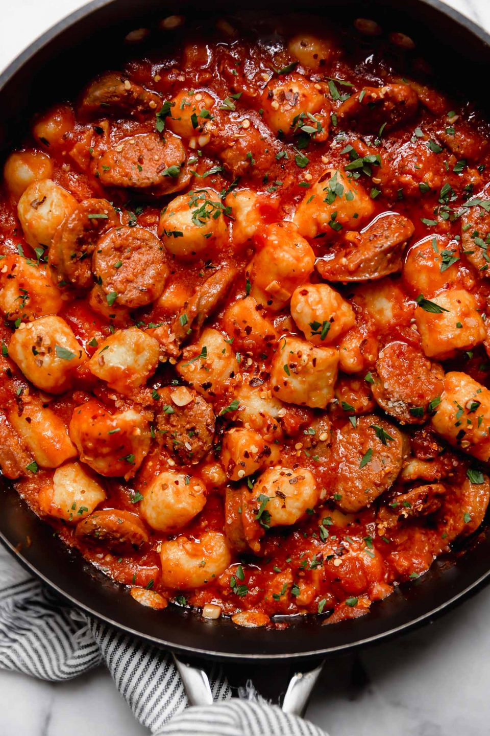 Gnocchi all'Amatriciana, with sliced Italian sausage in a black skillet. A striped linen is wrapped around the skillet handle. The skillet is on a white marble surface.