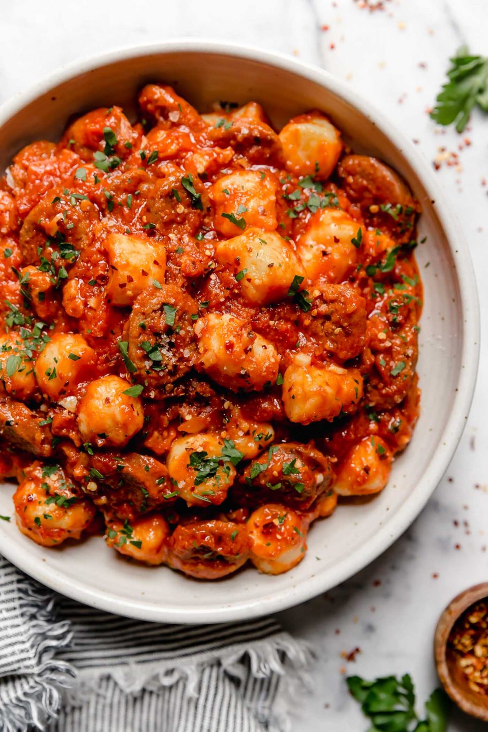 Gnocchi in Amatriciana sauce with Italian sausage. Served in a large white ceramic bowl, sitting on a white marble surface surrounded by fresh parsley.