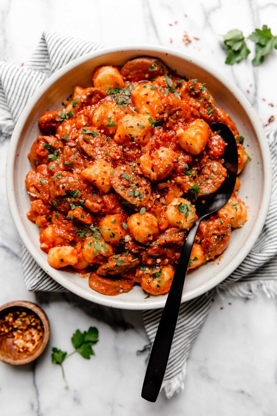 Gnocchi in Amatriciana sauce with Italian sausage. Served in a large white ceramic bowl with a large black serving spoon, sitting on a white marble surface surrounded by fresh parsley.