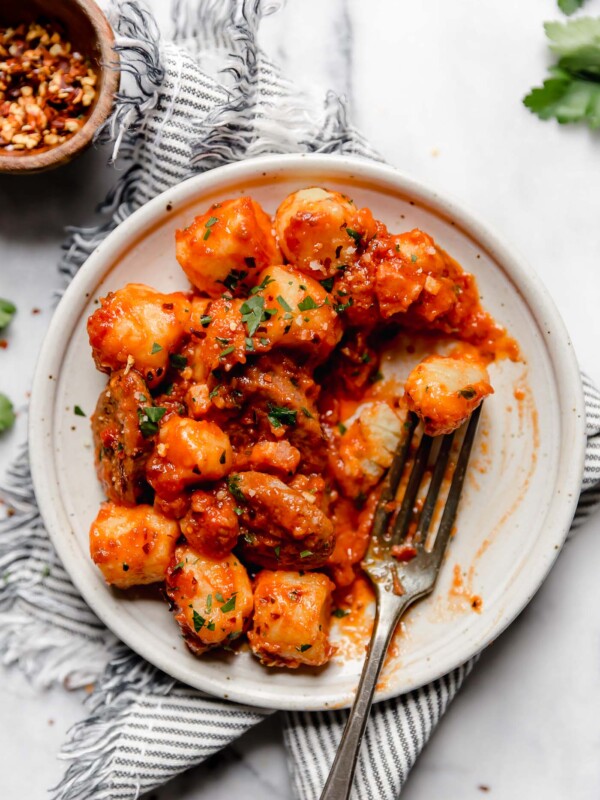 Gnocchi all'Amatriciana on a speckled white ceramic plate atop a striped linen napkin. A fork is in the gnocchi & amatriciana sauce, as though someone has been eating it. The plate is on a white marble surface, surrounded by fresh parsley leaves.