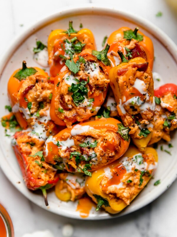 Buffalo chicken poppers on a small white ceramic plate. The poppers are topped with blue cheese dressing, buffalo sauce, & chopped green herbs. The plate is atop a white marble surface.