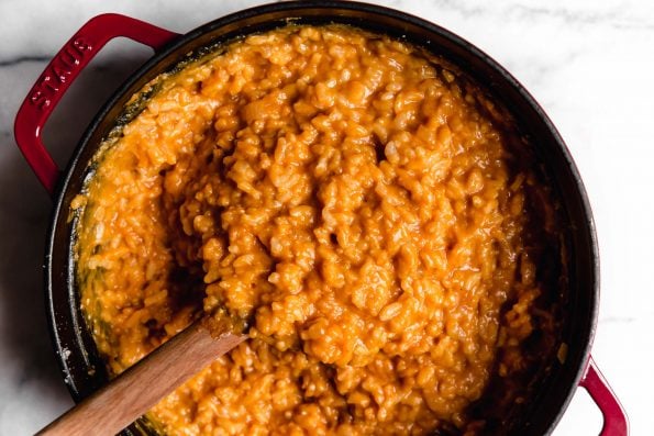 Creamy pumpkin risotto in a red Dutch oven. A wooden spoon holds up a spoonful of the risotto.