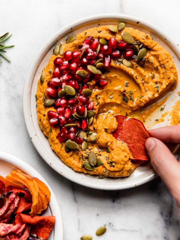 Creamy pumpkin hummus shown on a small speckled ceramic plate, topped with olive oil, pomegranate arils, pepitas, and fresh herbs. The hummus is placed on a white marble backdrop, next to some fresh rosemary and a plate of root veggie chips. A woman's hand is reaching into the plate, taking a scoop of it with a root veggie chip.