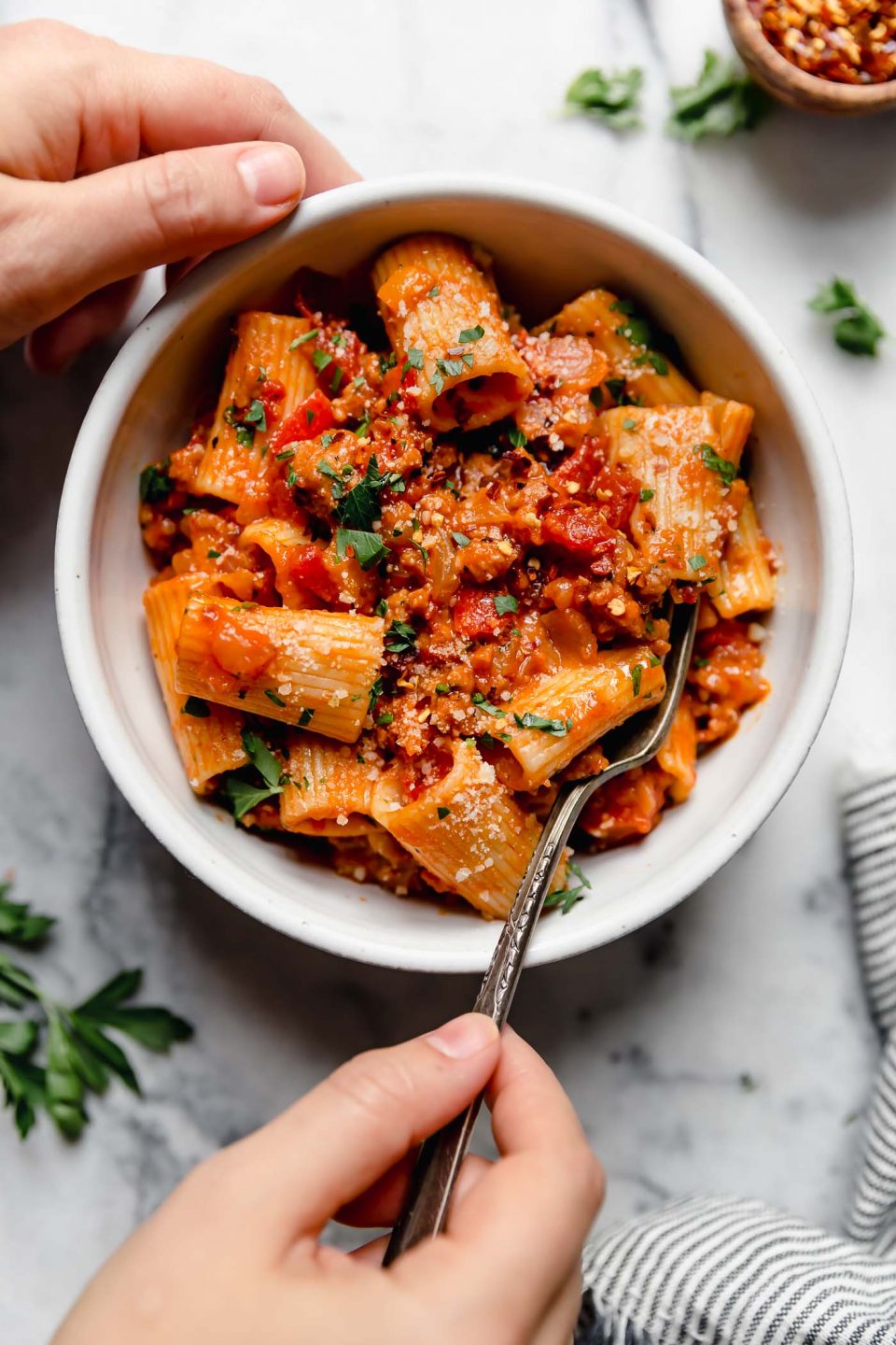 Italian Sausage & peppers pasta served in a white bowl sitting on a white marble surface surrounded by a striped linen napkin, fresh parsley leaves, & a small wooden bowl filled with crushed red pepper flakes.
