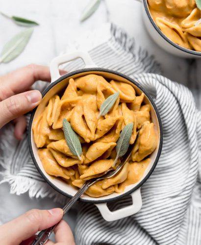 Vegan Pumpkin Mac and Cheese shown in a very small white ramekin. The dish is sitting on top of a gray + white striped napkin. The mac and cheese is topped with fresh sage leaves. A woman's hands are reaching into the frame of the photo with a fork, taking a bite out of the dish.