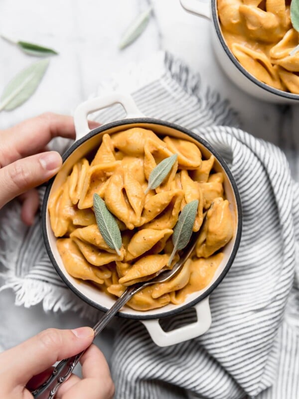 Vegan Pumpkin Mac and Cheese shown in a very small white ramekin. The dish is sitting on top of a gray + white striped napkin. The mac and cheese is topped with fresh sage leaves. A woman's hands are reaching into the frame of the photo with a fork, taking a bite out of the dish.